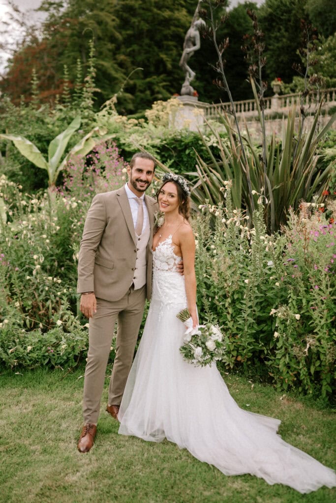 A newlywed couple poses in a lush garden at Port Lympne. The groom is dressed in a tan suit with a white shirt, while the bride wears a white gown with lace details, holding a bouquet of flowers. They stand close together, smiling, against a backdrop of greenery and colorful flowers—a moment beautifully captured by their Hythe wedding photographer. Image by Pearce Wedding Photography.