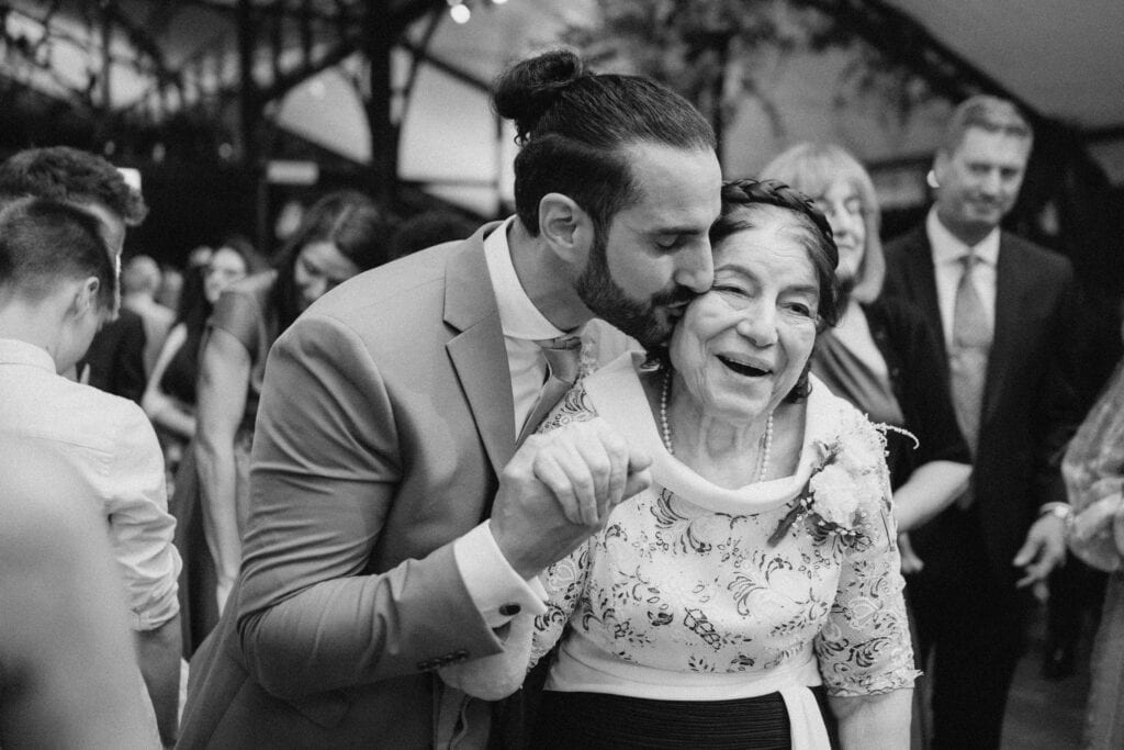 A man in a suit kisses the cheek of an older woman with braided hair and a floral dress as they smile and hold hands in a lively, crowded indoor setting. The black-and-white photo captures a warm, joyful moment, evoking the charm often seen in images by a Hythe wedding photographer. Image by Pearce Wedding Photography.