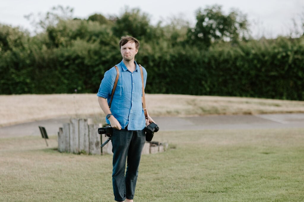 Man in blue shirt carrying cameras outdoors.