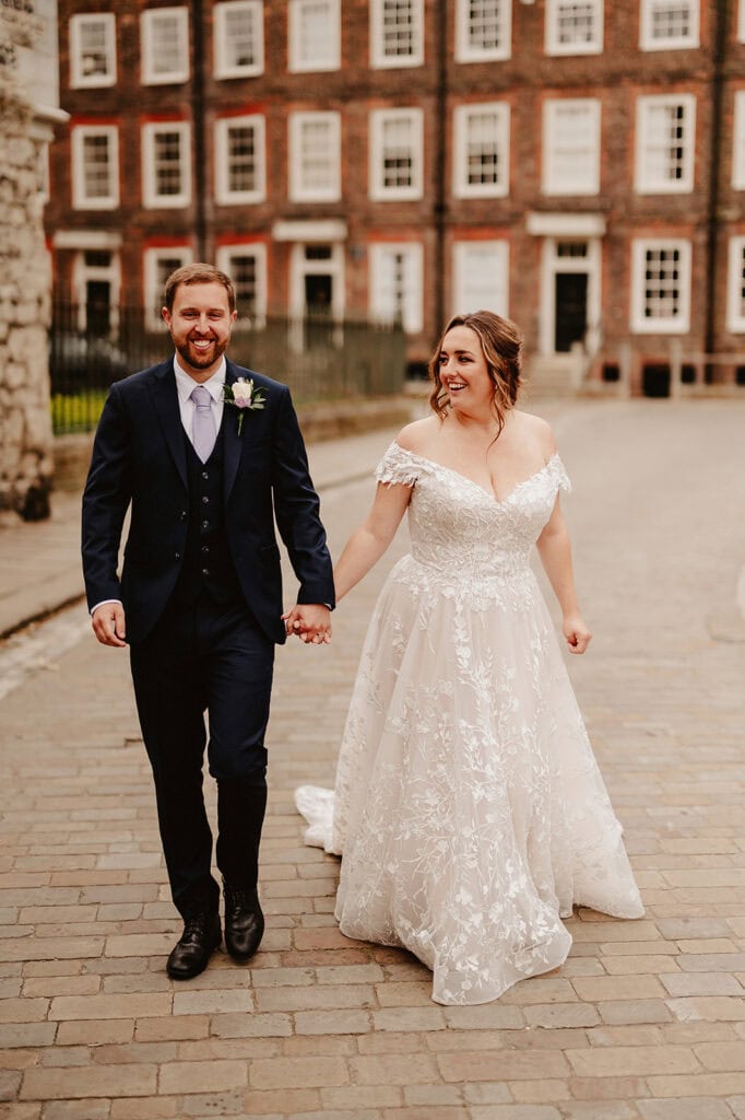 A couple holding hands and smiling, walking on a cobblestone street. The bride is in a white, off-shoulder lace wedding dress, and the groom is in a navy blue suit with a light purple tie and a white boutonniere. Captured by a Medway wedding photographer, they're framed by the historic buildings of The Vines of Rochester. Image by Pearce Wedding Photography.
