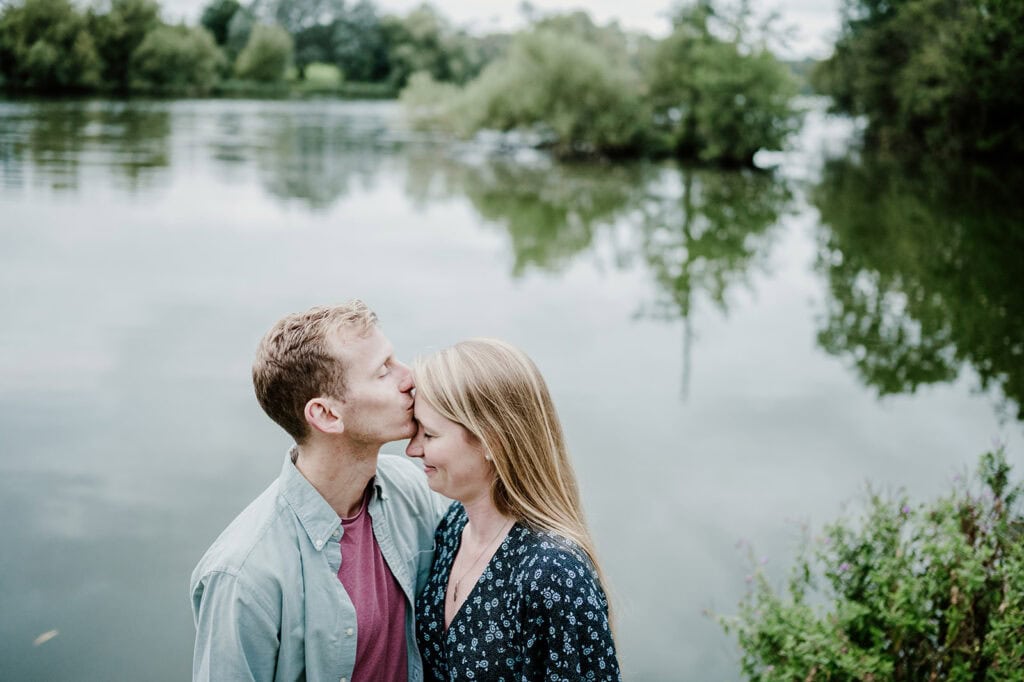 A man kisses a woman on the forehead as they embrace near a calm lake surrounded by greenery. The woman, smiling with her eyes closed, is clearly overjoyed at their recent engagement. Both are casually dressed, and the scene is serene with trees and water in the background under a cloudy sky. Image by Pearce Wedding Photography.
