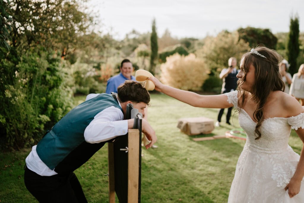 At The Gardens Yalding, a bride in a white wedding dress playfully pours a yellow substance over the groom's head as he bends over, leaning on a wooden structure. Guests smile and watch amidst the beautiful garden trees and shrubs. A moment of joy during their unforgettable wedding. Image by Pearce Wedding Photography.