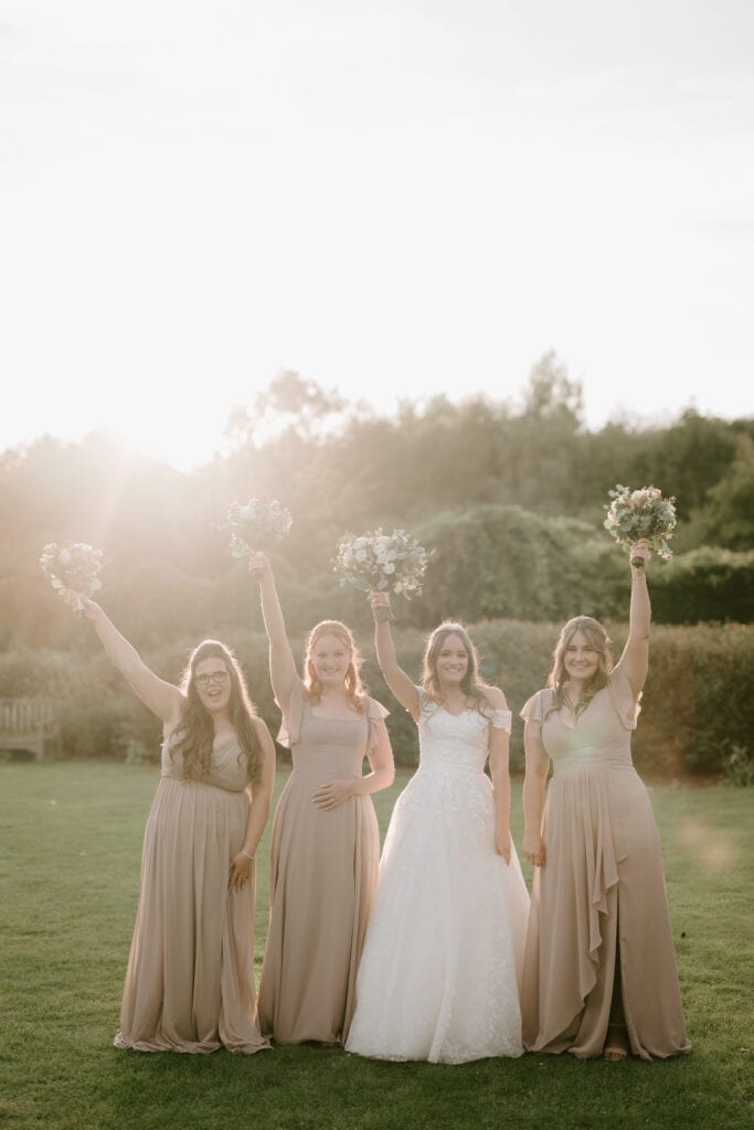 Four women stand on a lawn at The Gardens Yalding with sunlight in the background. The bride, in a white dress, is flanked by her bridesmaids wearing long, light-colored gowns. They all hold bouquets up high and smile warmly, celebrating the wedding occasion. Image by Pearce Wedding Photography.