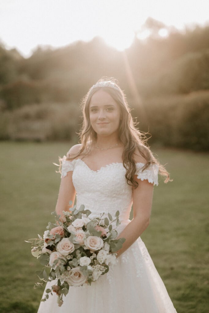 A bride stands outside in a lush green field at sunset, wearing an off-the-shoulder lace wedding dress from a renowned Kent bridal shop and holding a bouquet of white and pink flowers. Her long hair is loose, and she has a serene expression. The sun casts a warm glow behind her, creating a soft halo effect. Image by Pearce Wedding Photography.