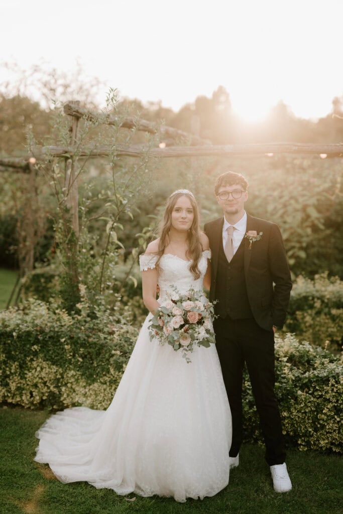 A bride and groom stand side by side in the enchanting gardens of Yalding during sunset. The bride, in a white off-shoulder wedding dress, holds a bouquet of flowers, while the groom is dressed in a black suit with a tie and boutonniere. The background features lush greenery and twinkling string lights. Image by Pearce Wedding Photography.