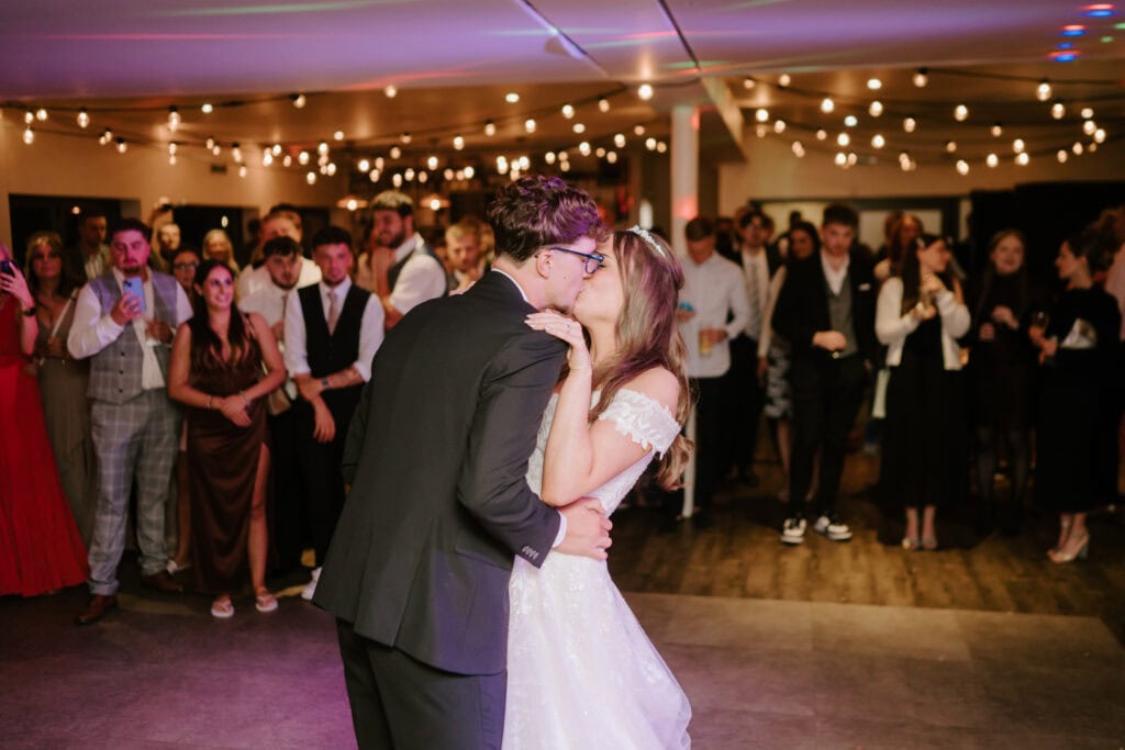 A newlywed couple shares a kiss on the dance floor during their wedding reception at The Gardens Yalding. The groom is in a black tuxedo and the bride is in a white gown. They are surrounded by guests, some applauding and others taking photos. String lights decorate the venue ceiling. Image by Pearce Wedding Photography.