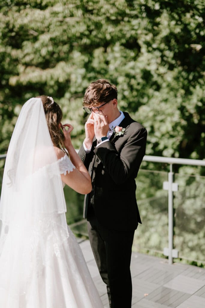 A bride and groom stand facing each other in the gardens Yalding. The bride, in a flowing white dress and veil, adjusts her veil while the groom, wearing a black suit and glasses, touches his face. The background is filled with lush green trees, setting a perfect wedding scene outdoors. Image by Pearce Wedding Photography.