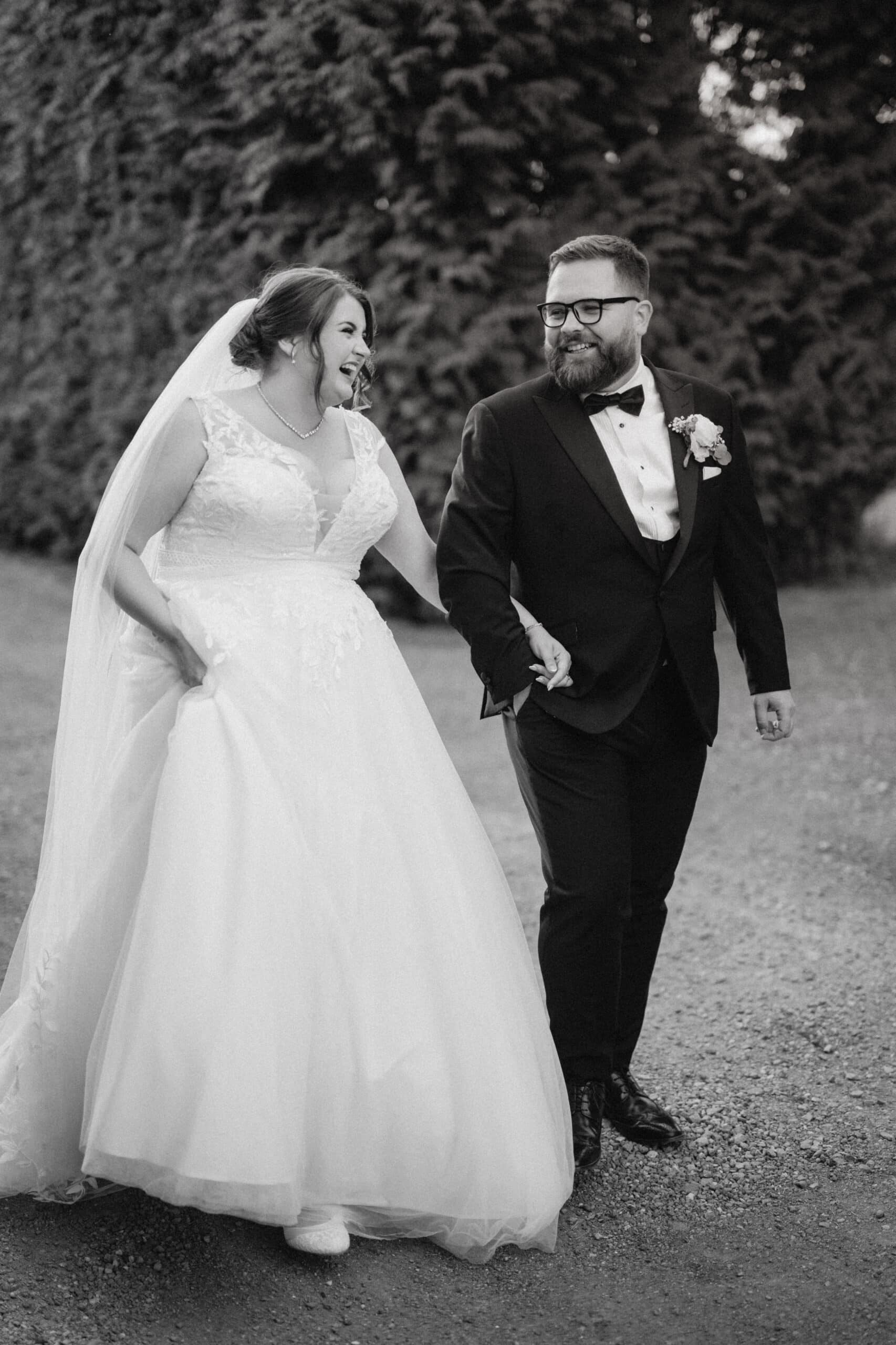 A bride in a white gown and veil and a groom in a black tuxedo walk hand in hand, smiling joyfully. Captured by a talented Kent wedding photographer, they stroll along a gravel path framed by tall greenery in the background. Image by Pearce Wedding Photography.