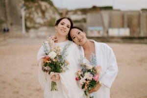 Two women in white outfits stand close together on a Broadstairs beach, smiling and holding colorful flower bouquets. The background reveals a sandy shore and distant buildings, perfectly captured by a Thanet wedding photographer. Image by Pearce Wedding Photography.