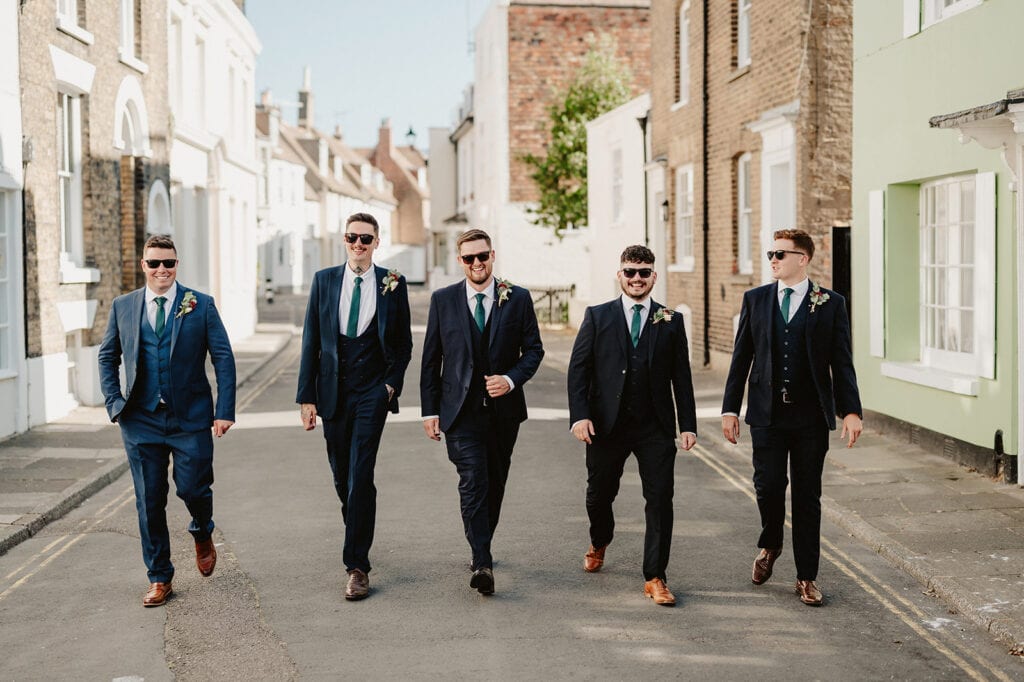 Five men in suits and sunglasses walk confidently down a sunny street, looking like they're striking a deal. They wear matching blue suits with green ties and boutonnieres, perfect for a wedding party. The background features charming brick and painted buildings. Image by Pearce Wedding Photography.