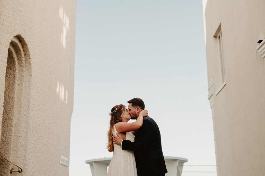 A newlywed couple shares a kiss between two light-colored buildings under a clear blue sky on the charming Deal seafront. The bride in her white dress and the groom in his dark suit complement the soft shadows falling across the textured walls, beautifully captured by their Deal wedding photographer. Image by Pearce Wedding Photography.