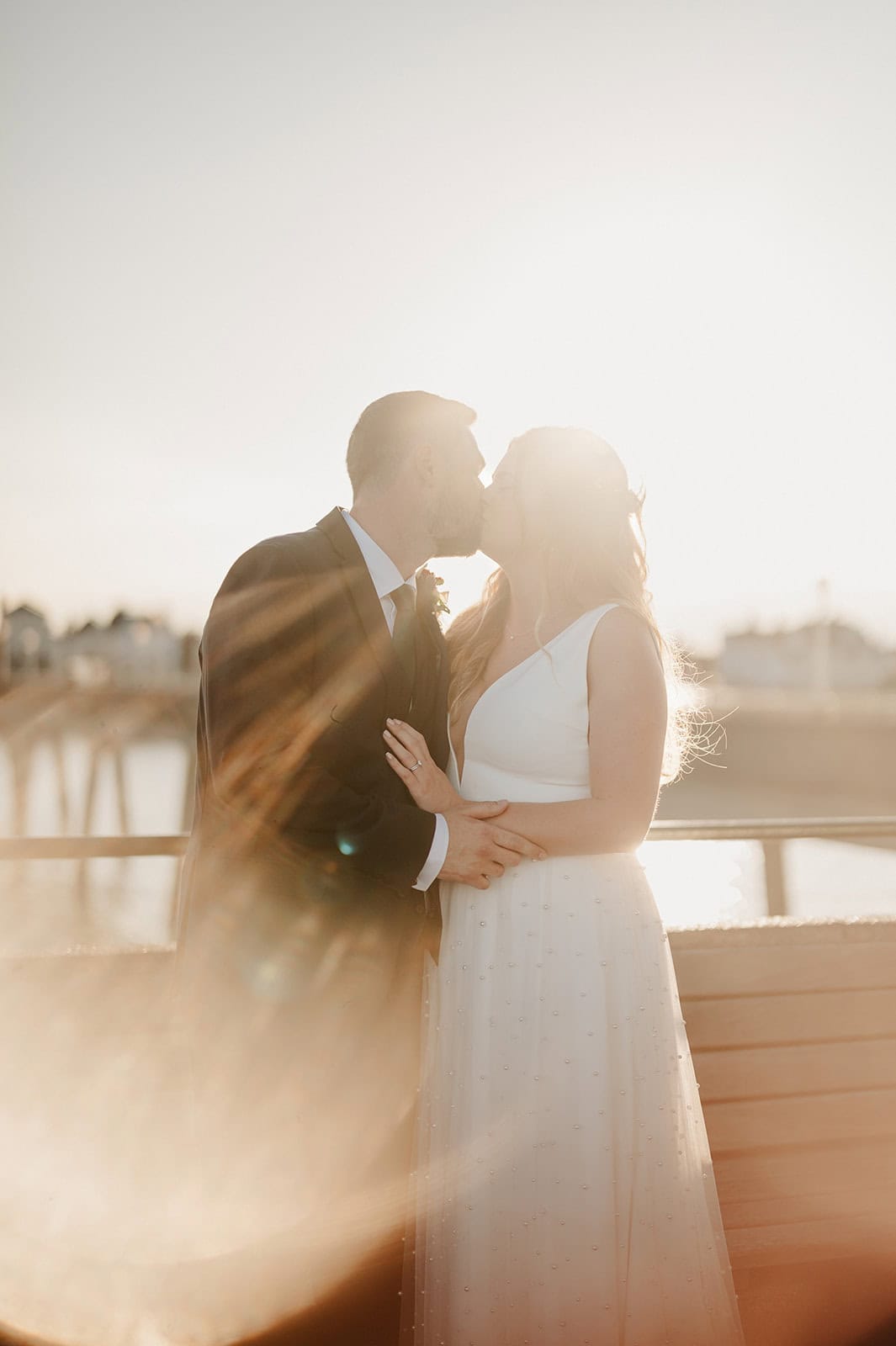 A couple shares a kiss at sunset on Deal Pier, the sun forming a warm halo around them. The bride's sleeveless white gown and the groom's dark suit add elegance. Blurred lights twinkle in the background, capturing a perfect wedding moment that's every photographer's dream. Image by Pearce Wedding Photography.