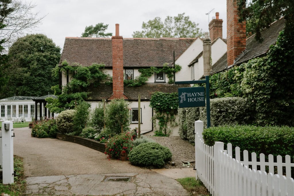 A charming, ivy-covered country house with a gabled roof and red brick chimneys stands behind a white picket fence. The sign reads "Hayne House," an enchanting venue in Kent. A garden path and lush greenery surround the house, while trees frame the picturesque backdrop perfect for weddings. Image by Pearce Wedding Photography.