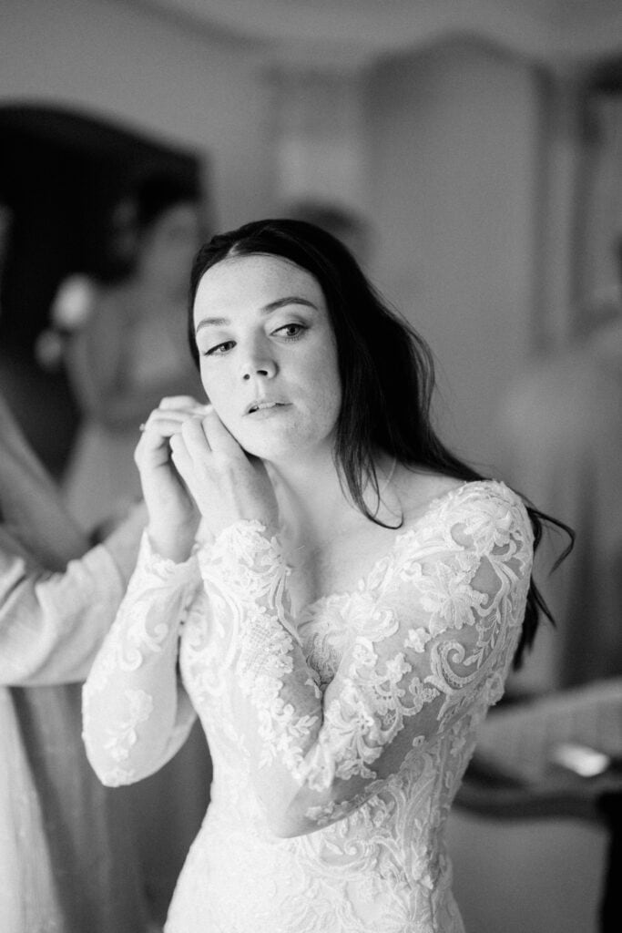 At the elegant Hayne House in Kent, a woman in a lace wedding dress adjusts her earring. Her long dark hair cascades as she looks slightly to the side. The softly blurred background focuses on her serene expression and detailed gown, capturing a perfect wedding moment. Image by Pearce Wedding Photography.