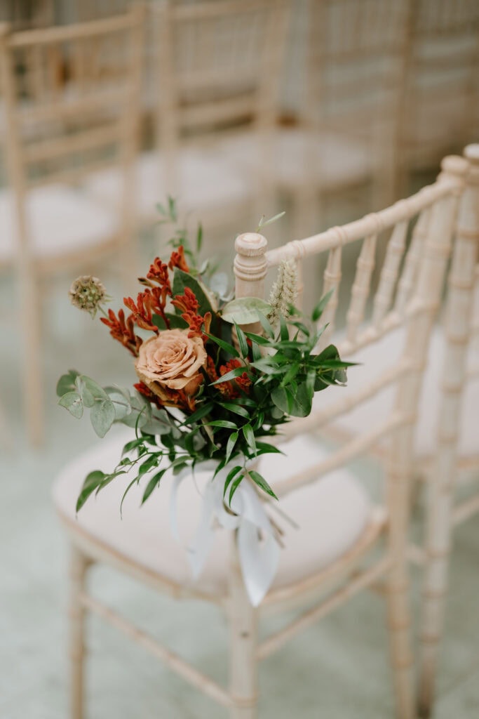 A bouquet with orange and red flowers, greenery, and a white ribbon adorns a light wood chair in a row of similar chairs, set up for a wedding at Hayne House in Kent. The setting exudes a serene and elegant atmosphere. Image by Pearce Wedding Photography.