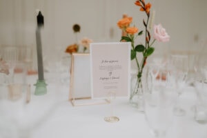 A table setting at Hayne House in Kent features a decorative card in a gold holder at the center, perfect for a wedding. The card boasts elegant text with vases of pink and orange flowers behind it. Clear wine and champagne glasses rest on the white tablecloth, completing the elegant scene. Image by Pearce Wedding Photography.