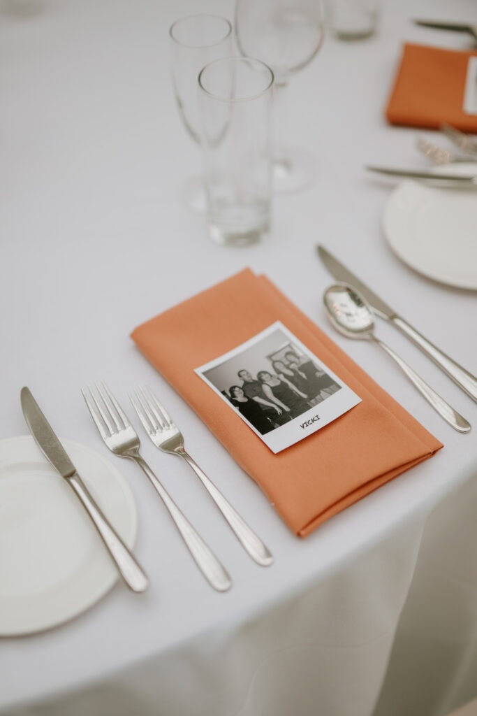 A formal table setting at Kent's Hayne House features a white tablecloth with a knife, fork, and spoon arranged neatly. An orange napkin holds a black-and-white wedding photo at its center. Two empty glasses are situated behind the napkin, creating an elegant atmosphere. Image by Pearce Wedding Photography.