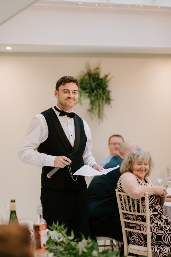At a wedding in Hayne House, a man in a black vest and bow tie stands smiling, holding a microphone and a piece of paper. He's speaking to guests seated at tables adorned with hanging greenery in the charming Kent setting, and everyone is listening intently with smiles. Image by Pearce Wedding Photography.
