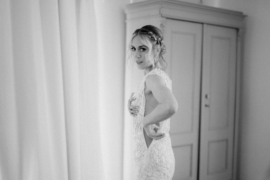 A black and white photo captures a serene moment of a person in a bridal gown, adjusting their dress near the light-colored curtain at Villiers Barn. Their braided hair complements the elegant atmosphere as they stand gracefully in front of the wardrobe, embodying Essex’s timeless wedding charm. Image by Pearce Wedding Photography.