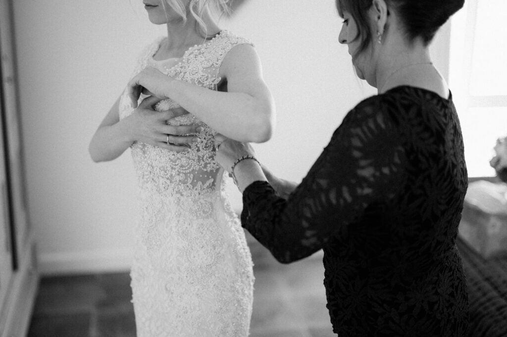 A black and white image of a bride in a detailed lace wedding dress, being assisted with zipping up the back, captures an intimate wedding moment indoors. The setting exudes charm reminiscent of Essex venues like Villiers Barn. Image by Pearce Wedding Photography.
