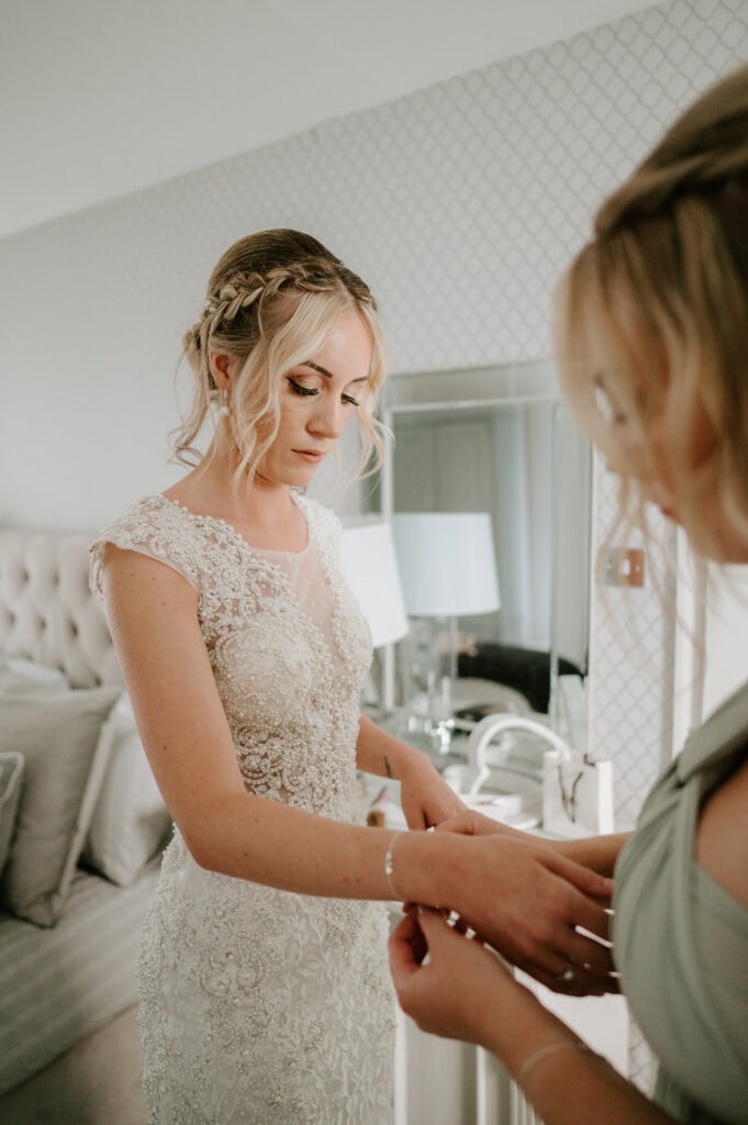 A bride in a lace wedding gown stands by a mirror at Villiers Barn, having a bracelet adjusted on her wrist by another person. The softly lit room, embodying modern decor, radiates an enchanting wedding atmosphere reminiscent of the charming Essex countryside. Image by Pearce Wedding Photography.
