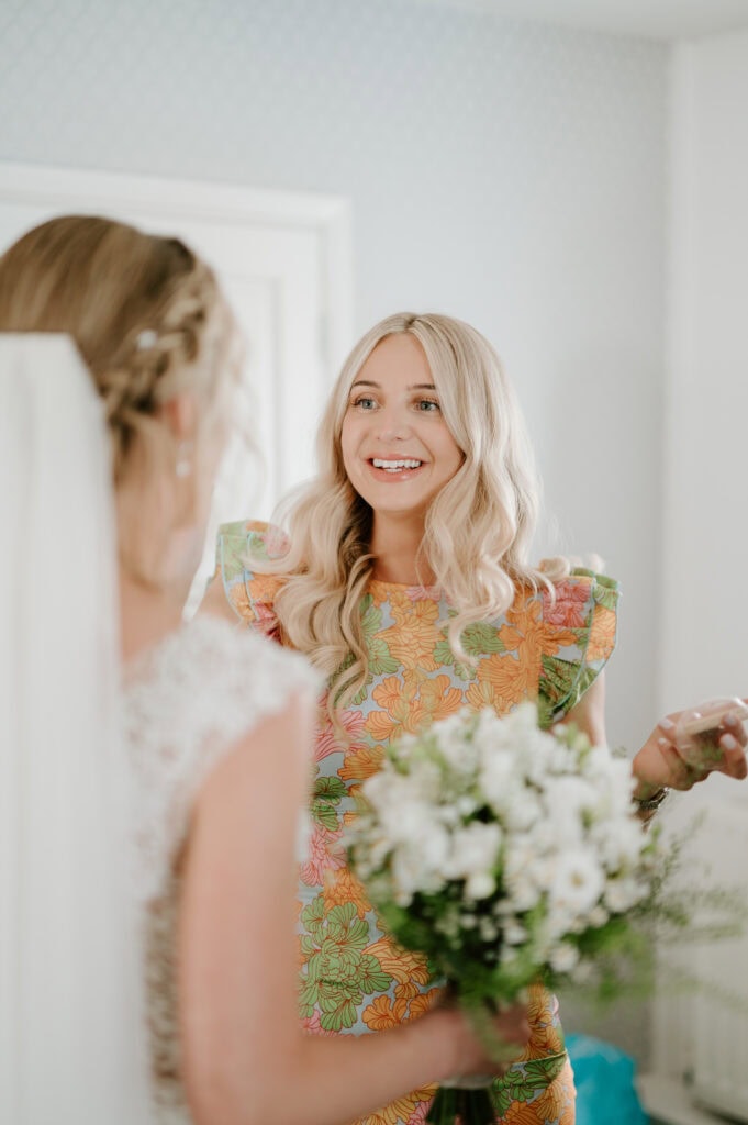 A woman with long blonde hair, wearing a colorful floral dress, smiles while holding a bouquet of white flowers. Another person with a veil is visible from behind, dressed in white, suggesting a wedding atmosphere. The room at Villiers Barn in Essex has light-colored walls. Image by Pearce Wedding Photography.
