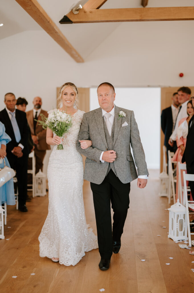 At Villiers Barn in Essex, a bride in a white lace dress holds a bouquet as she walks down the aisle arm-in-arm with an older man in a gray suit. Guests are seated along the aisle in the bright room adorned with rustic wooden beams, capturing the essence of this charming wedding setting. Image by Pearce Wedding Photography.