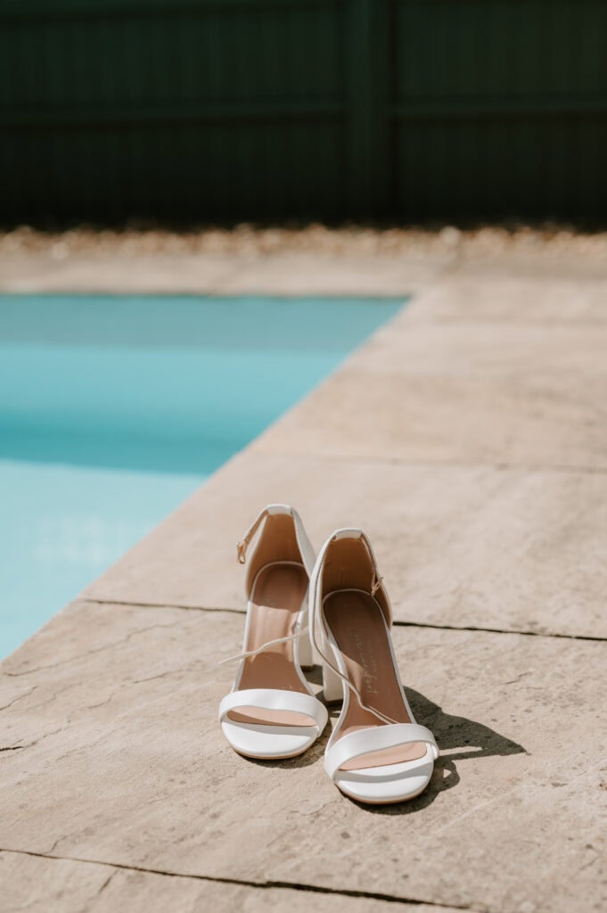 A pair of white, open-toed high heels rests on a stone surface beside a clear blue swimming pool at Villiers Barn. The sun casts soft shadows, hinting at a warm, sunny day perfect for a wedding in Essex. Image by Pearce Wedding Photography.