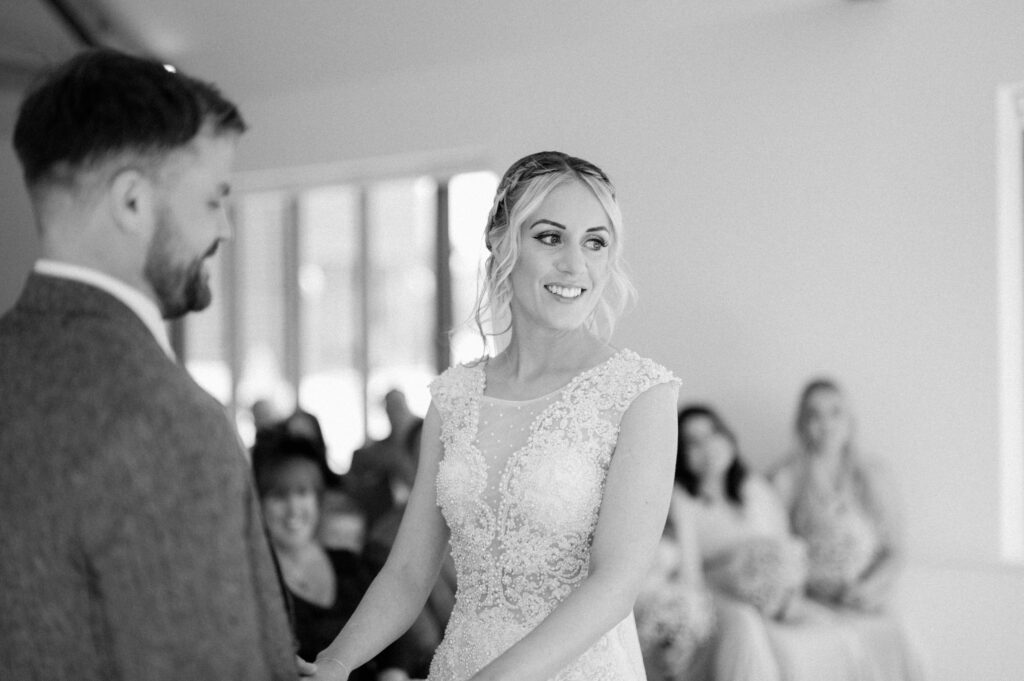 A black and white image captures a wedding at Villiers Barn in Essex. The bride, in a lace dress, smiles while holding hands with the groom in his suit. They stand before seated guests as soft lighting streams through large windows, creating an intimate atmosphere. Image by Pearce Wedding Photography.