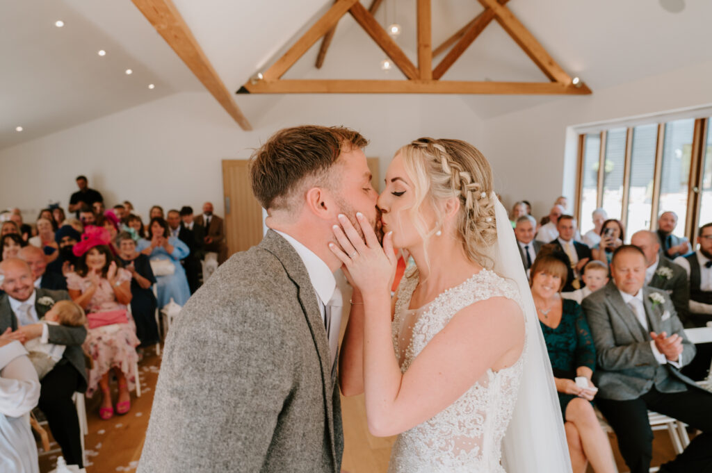 A bride and groom kiss during their wedding ceremony at Villiers Barn in Essex, surrounded by wooden beams. Guests sit in rows, joyfully watching the couple. The bride wears a lace gown and the groom dons a gray suit, creating an atmosphere that's both joyful and celebratory. Image by Pearce Wedding Photography.
