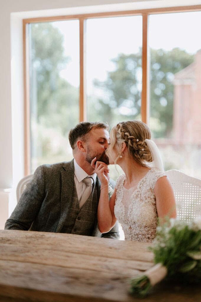 A bride and groom share a wedding kiss while seated at a wooden table in charming Villiers Barn. The groom dons a gray suit, and the bride is radiant in her lace dress with braided hair. A large window reveals the scenic beauty of Essex with its lush trees and distant buildings behind them. Image by Pearce Wedding Photography.