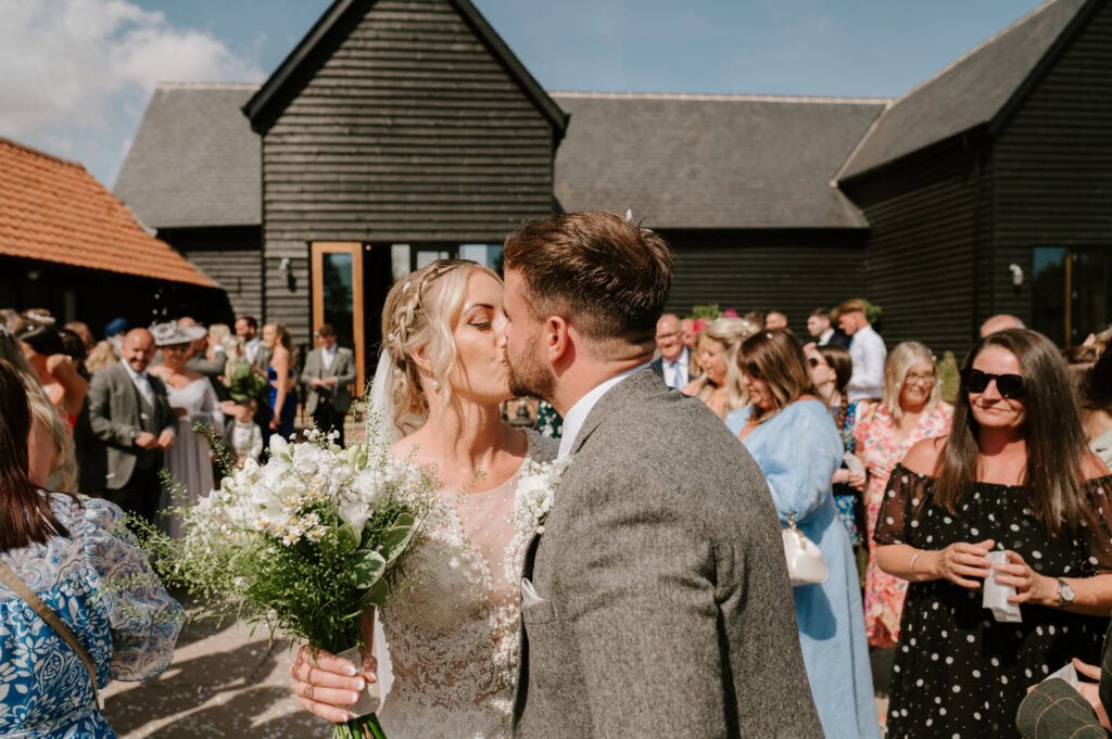 A newlywed couple kisses outdoors in front of a crowd at Villiers Barn during their Essex wedding. The bride holds a bouquet of white flowers and wears a lace dress, while the groom is in a gray suit. Guests smile and enjoy the celebration under the sunny sky. Image by Pearce Wedding Photography.