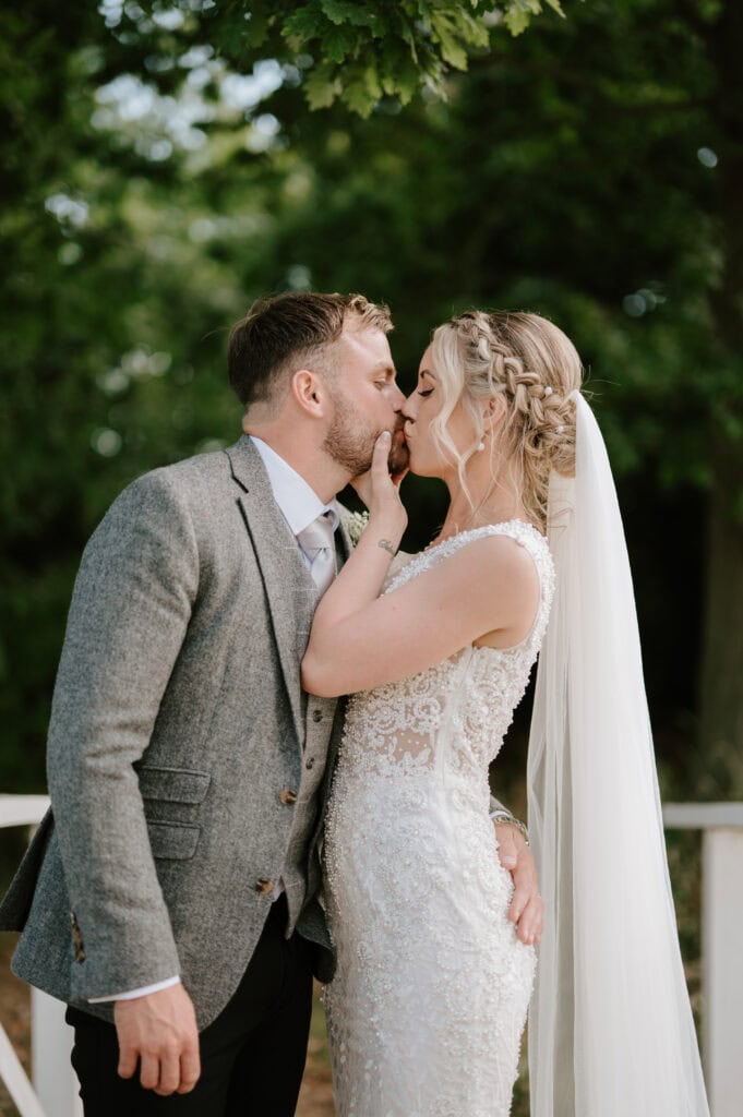 A couple kisses outdoors, surrounded by lush greenery at Villiers Barn in Essex. The woman stuns in a lace wedding dress with a long veil, while the man looks sharp in a gray suit. They stand on a wooden bridge, embracing the romantic and intimate moment of their wedding day. Image by Pearce Wedding Photography.