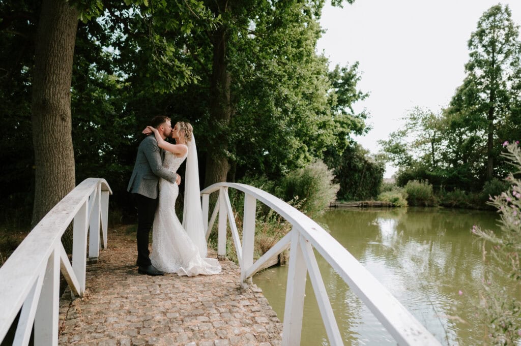 A bride and groom embrace on a small arched bridge over a pond at Villiers Barn in Essex, surrounded by lush green trees. The bride wears a long white gown and veil, and the groom is in a dark suit. The scene is peaceful and romantic. Image by Pearce Wedding Photography.