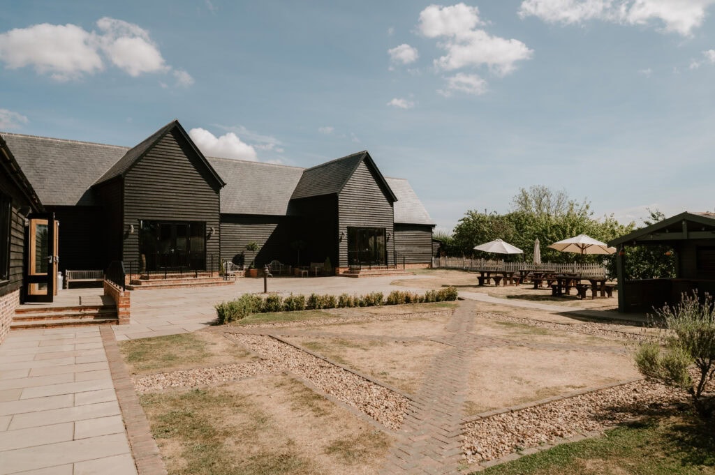 Villiers Barn in Essex, a rustic wooden building with a grey roof, sits under a blue sky with scattered clouds. The foreground features a paved patio and picnic tables with umbrellas, perfect for hosting an intimate wedding. Trees and bushes provide a charming backdrop. Image by Pearce Wedding Photography.