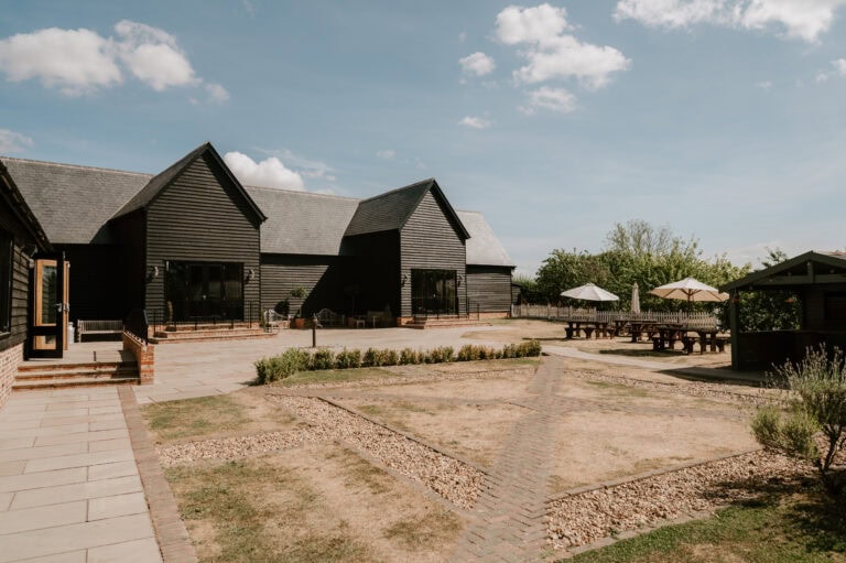 Villiers Barn in Essex, a rustic wooden building with a grey roof, sits under a blue sky with scattered clouds. The foreground features a paved patio and picnic tables with umbrellas, perfect for hosting an intimate wedding. Trees and bushes provide a charming backdrop. Image by Pearce Wedding Photography.