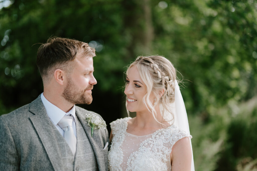 Amidst the lush greenery of Villiers Barn in Essex, a bride and groom gaze lovingly at each other. The bride’s lace gown and braided hair complement the groom’s gray suit and boutonniere perfectly, capturing a timeless wedding moment outdoors. Image by Pearce Wedding Photography.