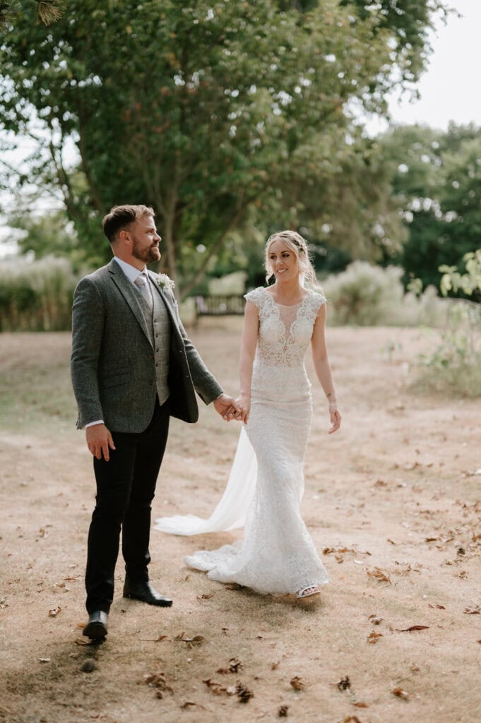 A couple holding hands strolls through a sunlit, grassy area at Villiers Barn in Essex. The bride wears a lace wedding dress with a long train, while the groom sports a grey suit with a vest. Green trees and the beautiful outdoor scenery create the perfect backdrop for their wedding day. Image by Pearce Wedding Photography.