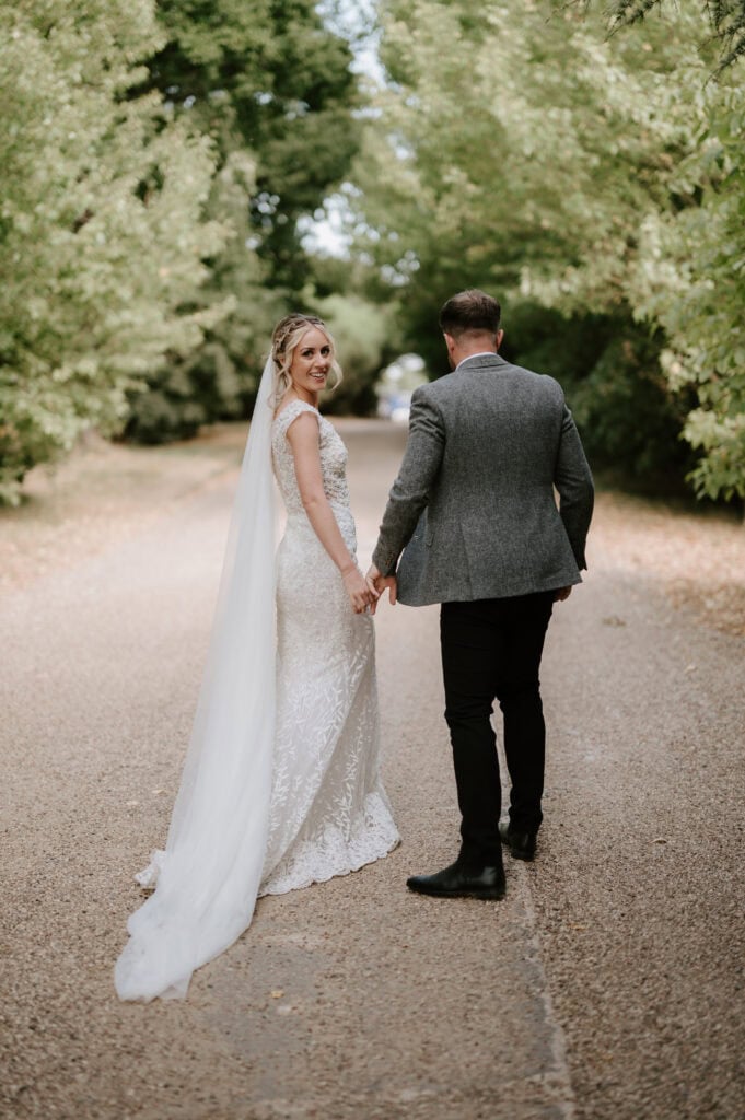 A bride and groom are walking hand in hand down a tree-lined path at Villiers Barn. The bride, in a stunning white gown with a veil, and the groom, elegantly dressed in a gray suit with black pants, glance back towards the camera amidst Essex's lush greenery. Image by Pearce Wedding Photography.