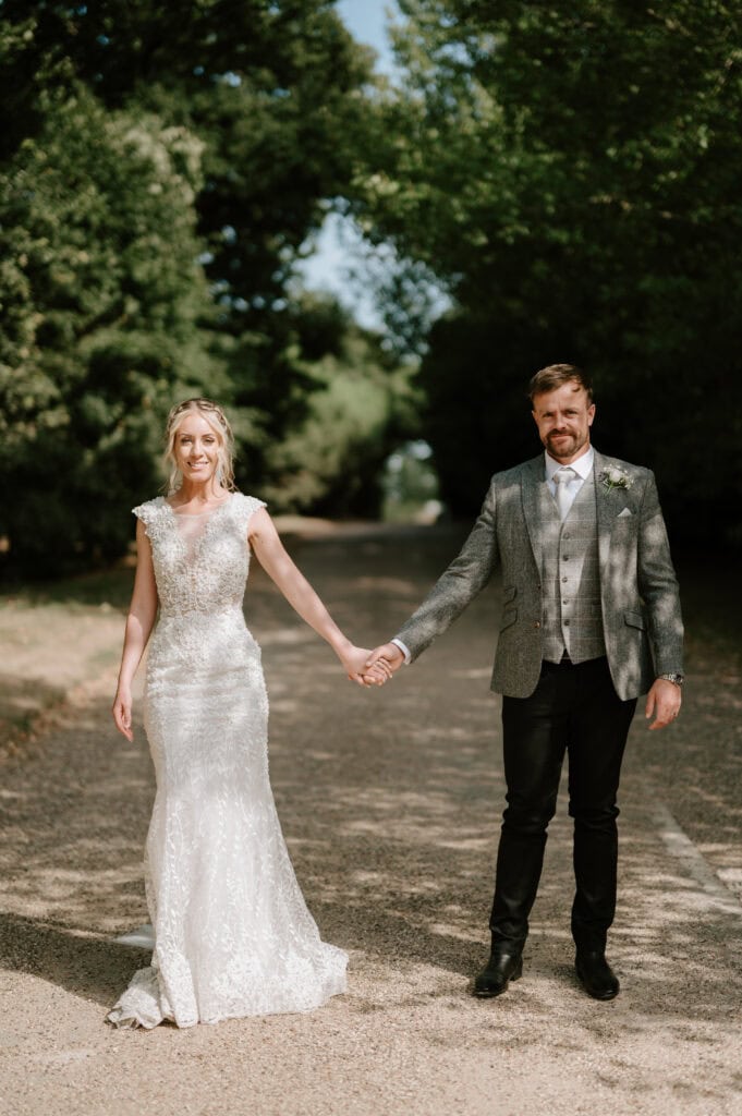 A bride and groom hold hands while standing on a tree-lined path at Villiers Barn in Essex. The bride is wearing a white lace gown, and the groom is in a gray jacket with a vest and tie. Sunlight filters through the trees, casting a warm glow on the couple, capturing their wedding day bliss. Image by Pearce Wedding Photography.
