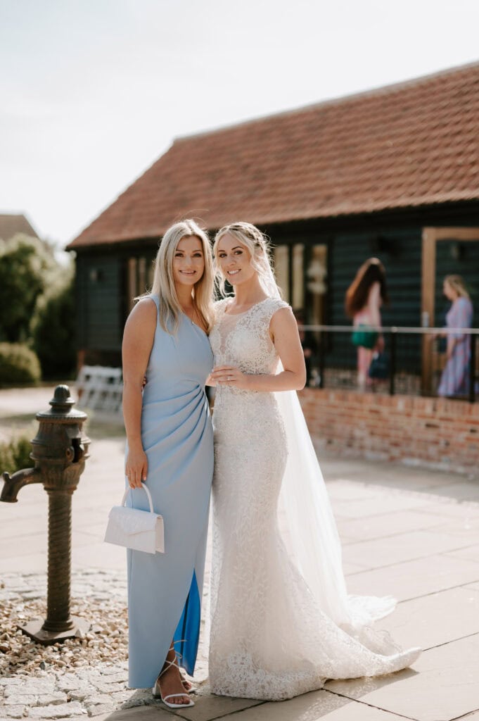 Two women smiling outside in front of Villiers Barn. One wears a light blue dress and holds a white purse, while the other is adorned in a white lace wedding gown with a veil, embodying the perfect Essex wedding day charm. Image by Pearce Wedding Photography.
