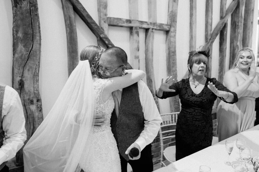 In the charming ambiance of Villiers Barn in Essex, a bride in a wedding dress and veil hugs an older man in a vest. Nearby, a woman in a dark dress and hat gestures animatedly, while another woman in a light dress claps her hands amidst the rustic wooden beams. Image by Pearce Wedding Photography.