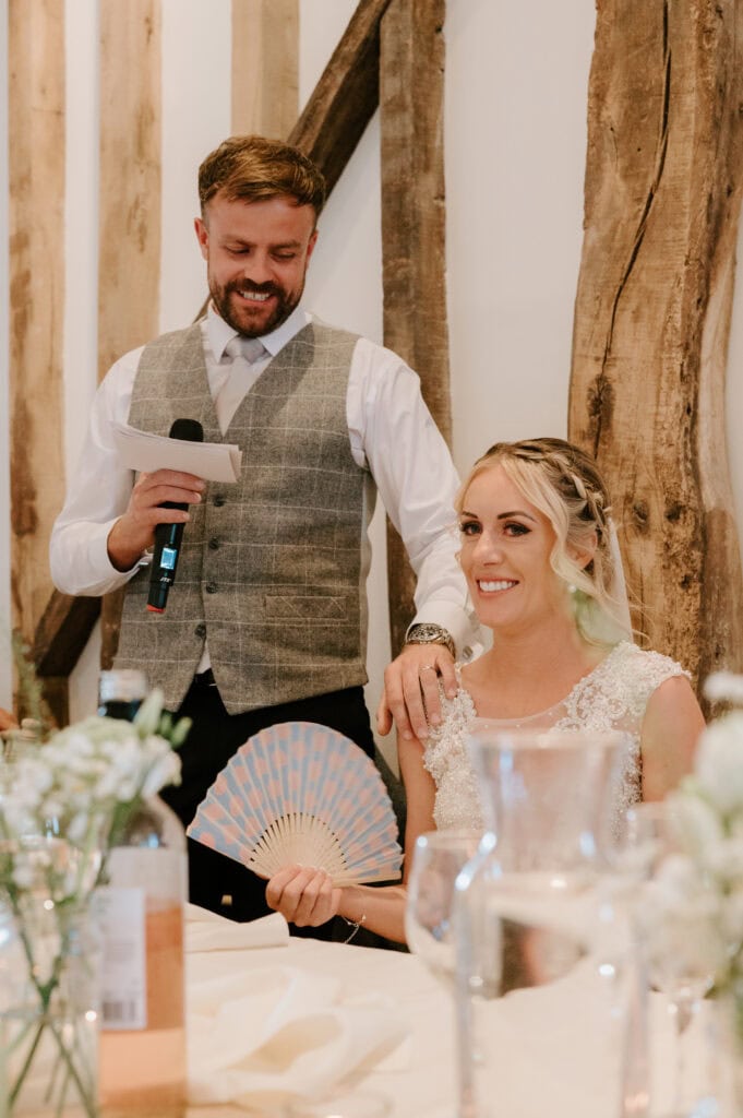 A man in a vest holds a microphone and a piece of paper, standing beside a seated woman in a wedding dress, who is smiling and holding a fan. They are indoors at Villiers Barn, with rustic wooden beams in the background, capturing the charm of an Essex wedding scene. Image by Pearce Wedding Photography.
