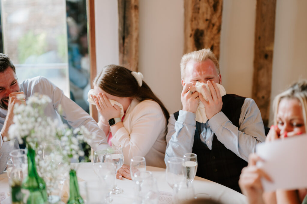 Three people sit at a round table with a white tablecloth, holding napkins to their faces as if wiping away tears of laughter. The table is adorned with glasses, bottles, and a floral centerpiece. Wooden beams hint at the rustic charm of Villiers Barn in Essex, creating an idyllic wedding scene. Image by Pearce Wedding Photography.