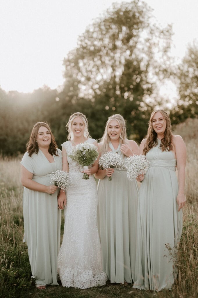 Four women stand outdoors in a grassy field at Villiers Barn. The bride, in a white lace gown holding flowers, is flanked by three bridesmaids in matching light green dresses. They smile warmly against an Essex backdrop of trees with sunlight peeking through. Image by Pearce Wedding Photography.