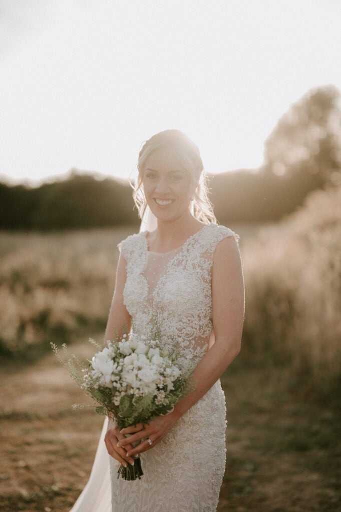 In the serene landscape of Essex, a bride in a detailed lace wedding dress holds a bouquet of white flowers. Standing in the sunlit, grassy field near Villiers Barn, she smiles with trees in the background, capturing the essence of a perfect wedding day. Image by Pearce Wedding Photography.