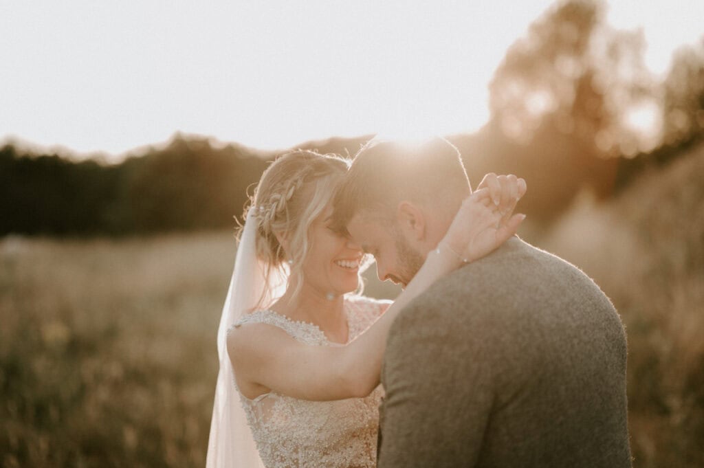 A couple embraces in a field at sunset outside Villiers Barn. The woman, wearing a veil and wedding dress, holds the man's face as they smile tenderly. Sunlight filters through the trees, casting a warm and romantic Essex glow over their wedding day moment. Image by Pearce Wedding Photography.