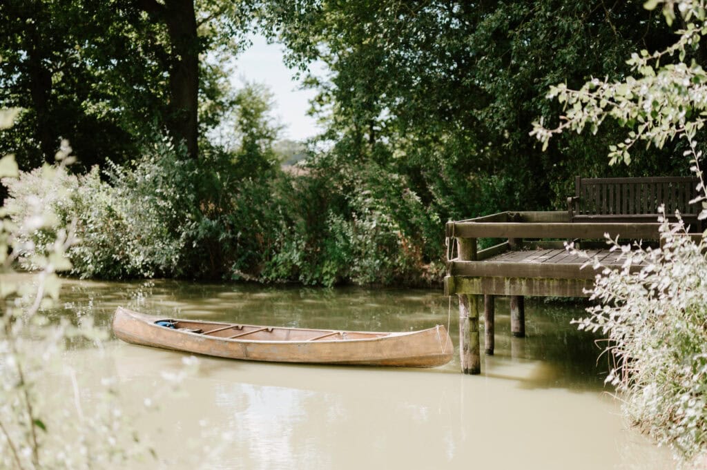 A wooden canoe floats on a calm, green-tinted river beside a rustic dock surrounded by the lush greenery and trees of Villiers Barn in Essex, creating a perfect backdrop for a sunny wedding day. Image by Pearce Wedding Photography.