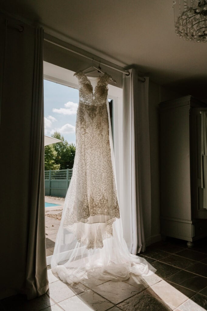 A white lace wedding dress hangs gracefully in a sunlit doorway at Villiers Barn, Essex. The intricate embroidery and long, sheer train catch the light beautifully, casting soft shadows on the floor. Outside, the garden blooms beside a serene pool. Image by Pearce Wedding Photography.