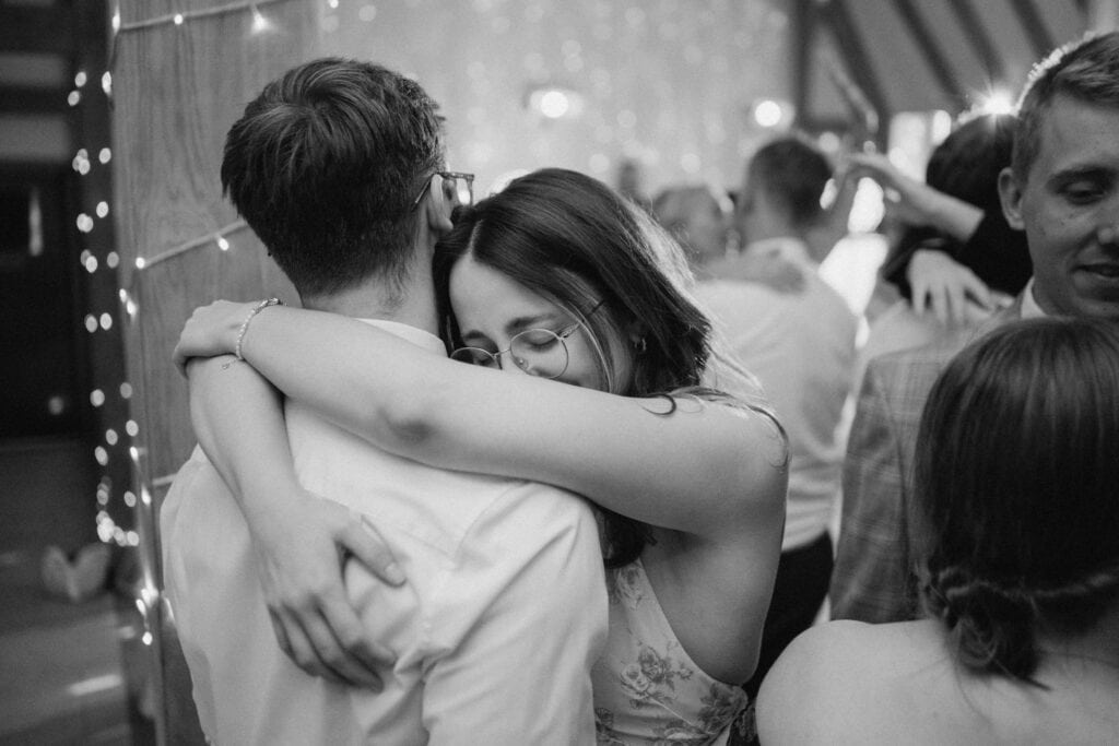 A black and white photo captured by a Kent wedding photographer shows a couple embracing tightly in a warm hug on the dance floor. The setting is festive, with blurred lights and joyful guests dancing in the background. The woman wears glasses and smiles softly, radiating happiness and affection. Image by Pearce Wedding Photography.